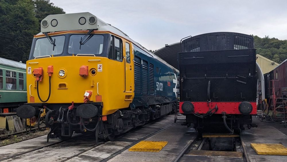 Two trains parked on tracks outside a shed, one yellow and blue, the other black with red detailing.
