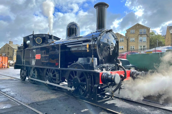 Black steam locomotive emits steam on a sunny day with buildings in background.