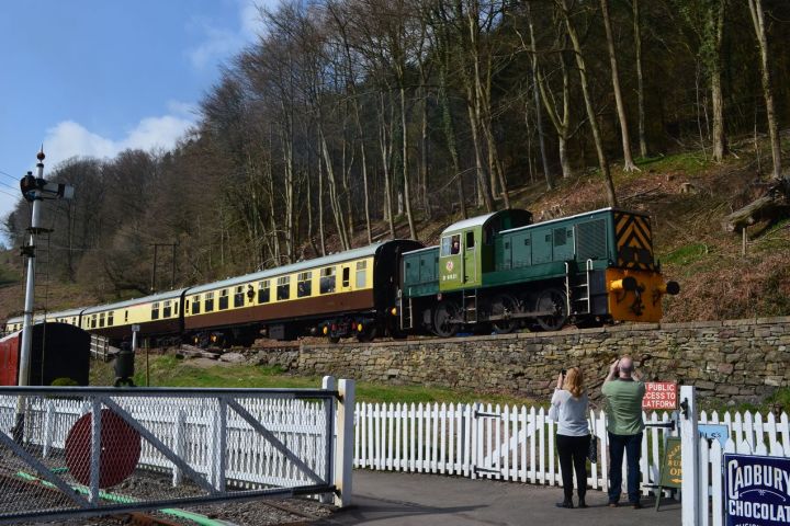 Vintage train on tracks with green locomotive and yellow carriages, people watching, trees and blue sky in background.