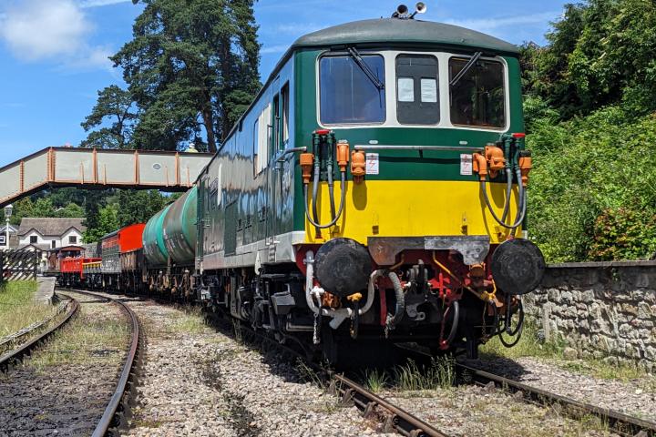 Colorful freight train on a rural track under a blue sky, with trees and a footbridge.
