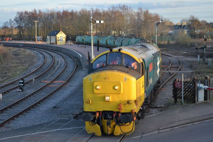 Yellow locomotive on railway tracks, pulling green tankers in a rural setting.