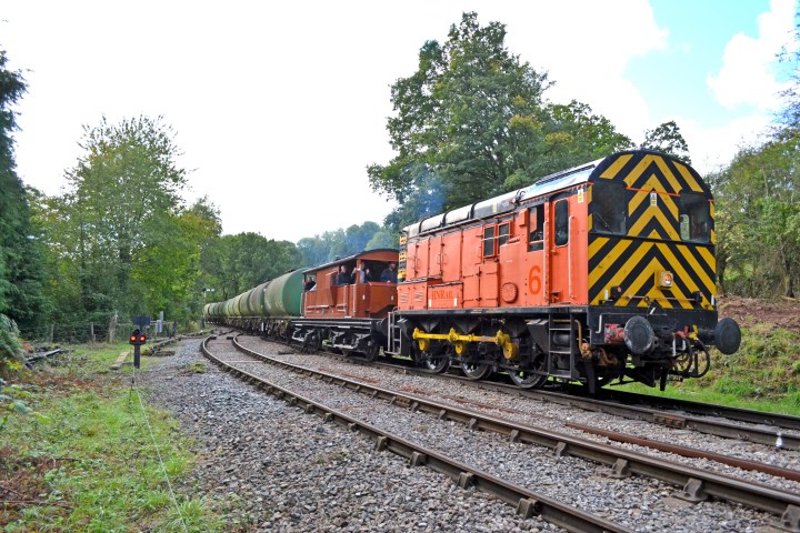 Orange train engine pulling tankers on a railroad track surrounded by trees.