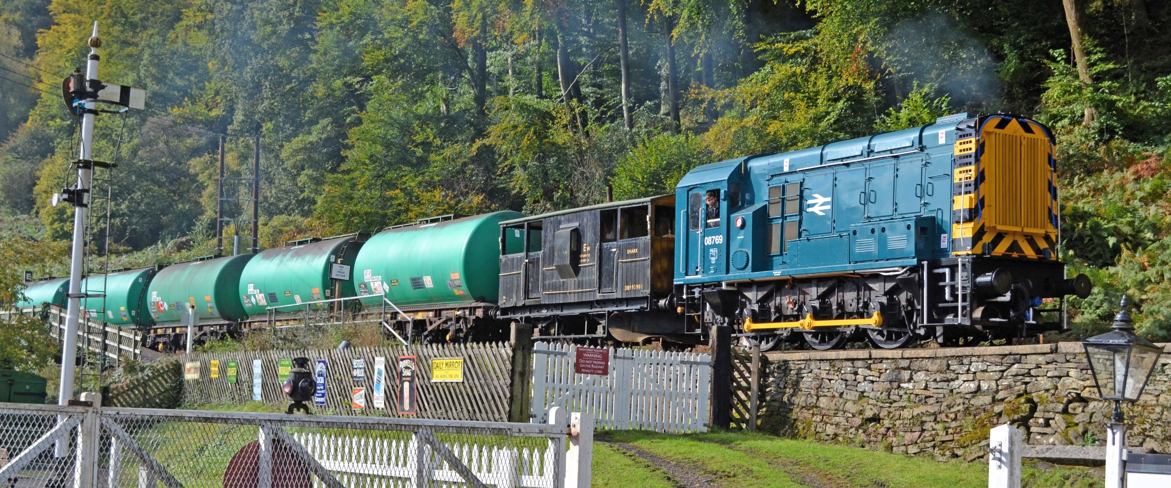 Blue diesel train with green tank cars on a track near a forested hillside.