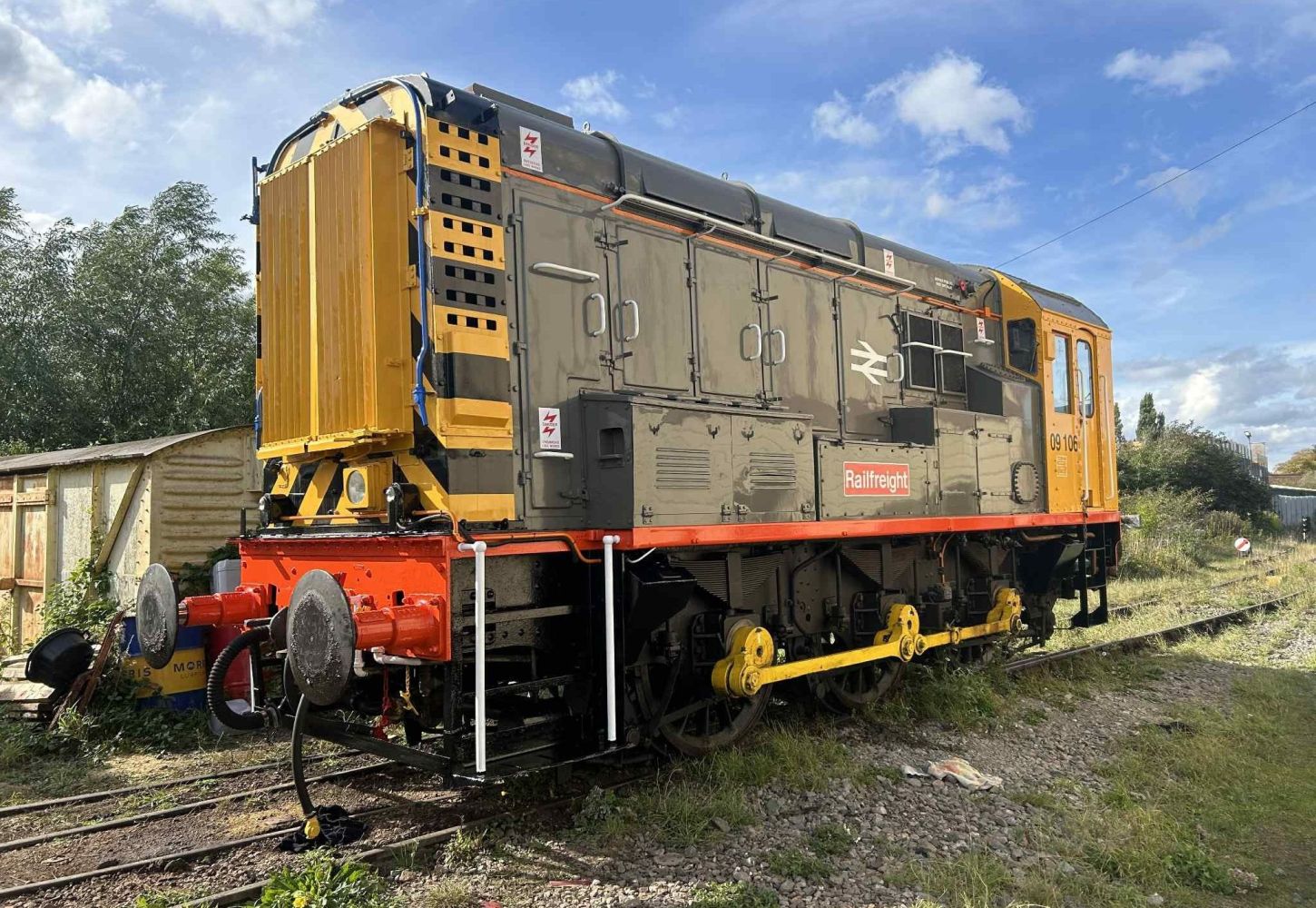 Yellow and gray diesel locomotive on railway tracks with trees and shed in background.