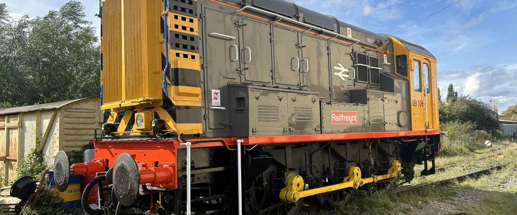 Yellow and gray diesel locomotive on railway tracks with trees and shed in background.