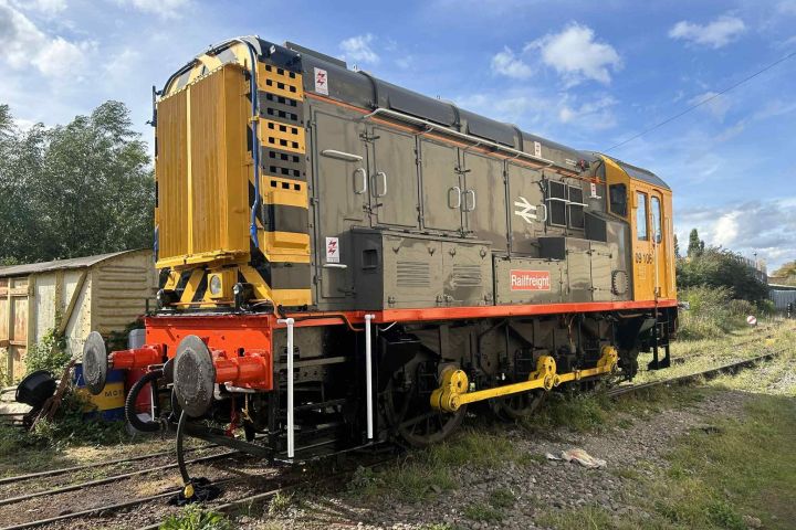 Yellow and gray diesel locomotive on railway tracks with trees and shed in background.