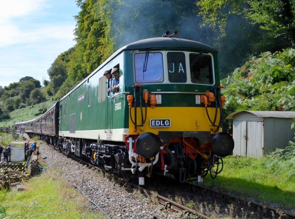 Green train with people inside on a rural track with trees and shed nearby.