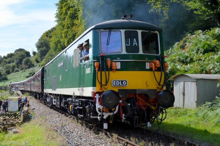 Green train with people inside on a rural track with trees and shed nearby.