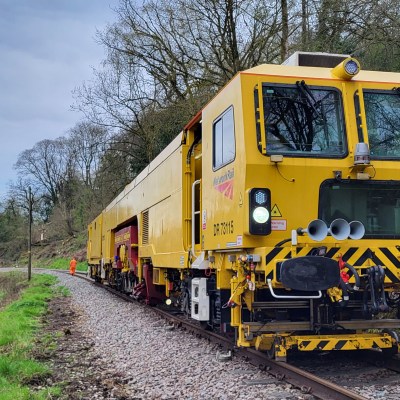Yellow rail maintenance train on tracks near wooded area with overcast sky.