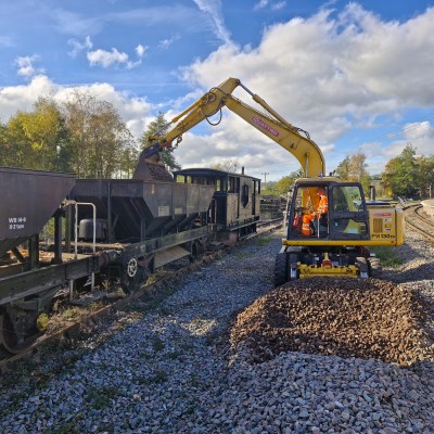 Excavator unloading gravel onto railroad tracks beside a train and station.