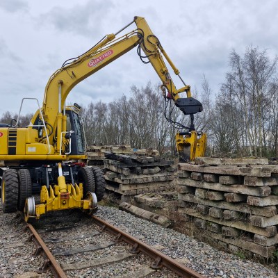 Yellow excavator on railroad tracks lifting concrete sleepers near trees.