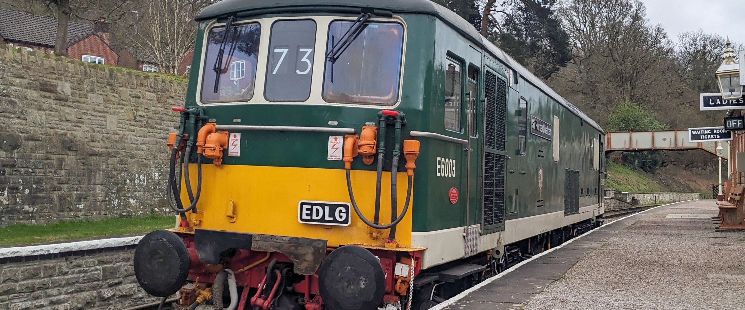 Green and yellow vintage train stopped at a station platform with trees in the background.