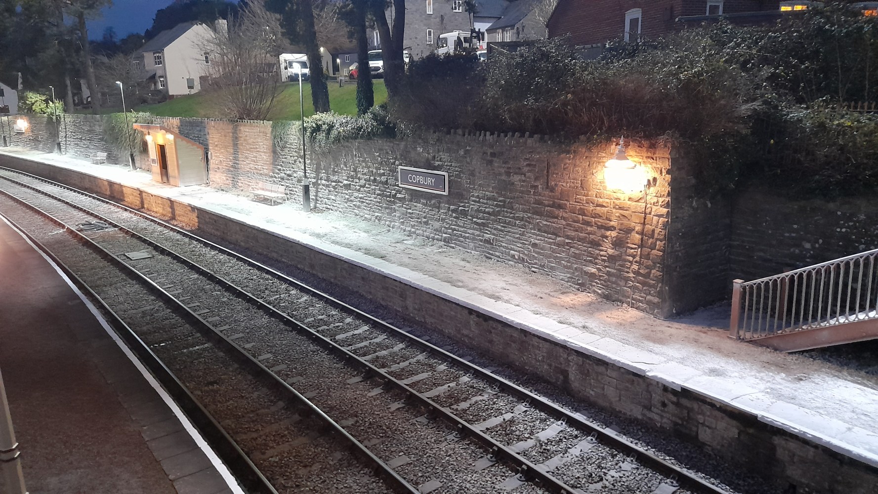 Empty train platform at night with dim lighting and buildings in background.