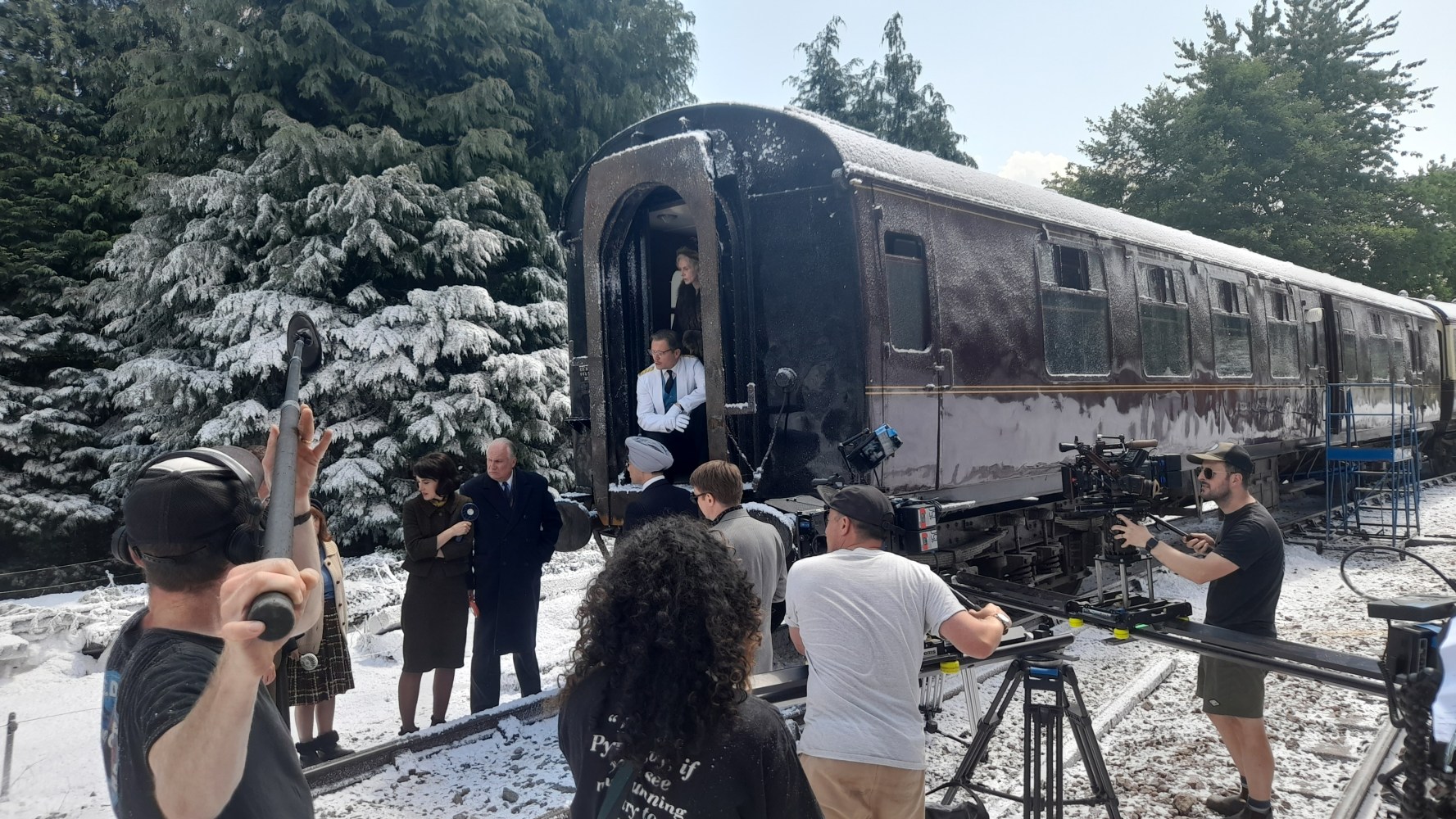Film crew shooting a scene by a vintage train with actors in period costumes and snow-covered scenery.