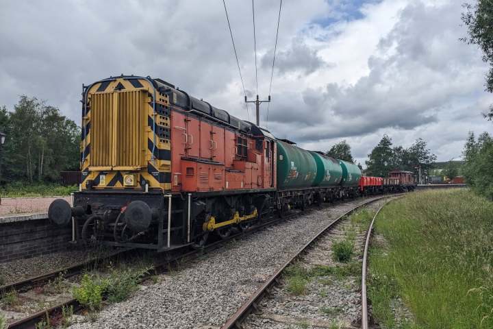 Red train with green tank cars on railway tracks under cloudy sky.
